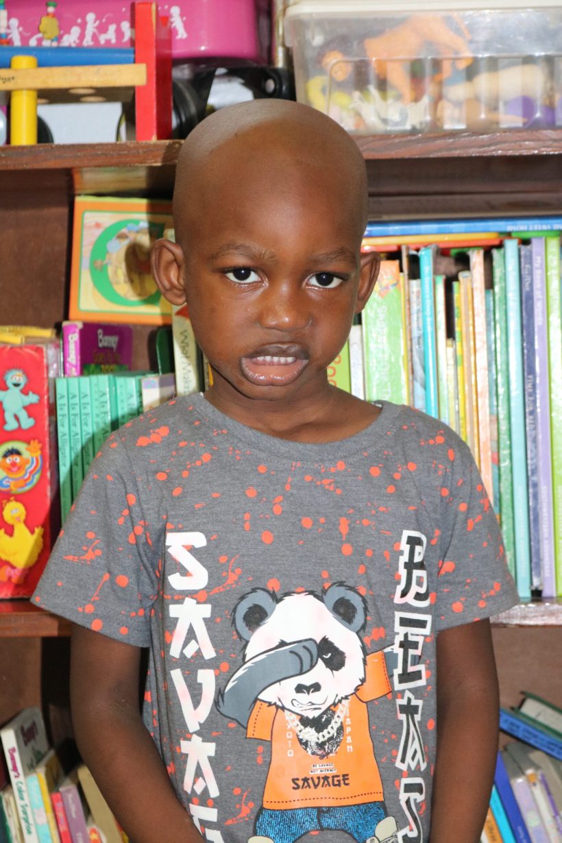 A preschool boy in a graphic t-shirt in front of the classroom bookshelves
